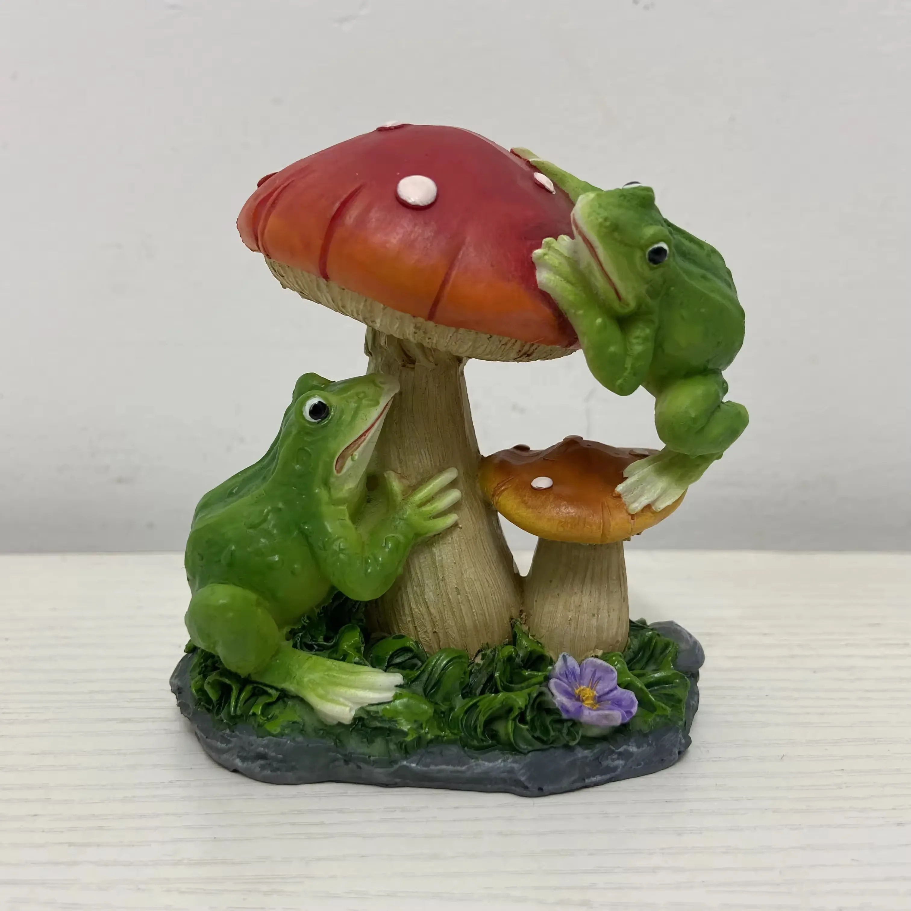 Dos figurines de rana en resina trepando alrededor de hongos rojos y naranjas en una escena de jardin divertida