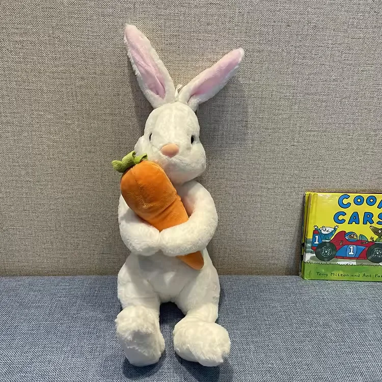 Peluche de conejo blanco con interior de orejas rosa abrazando una zanahoria de gran tamaño