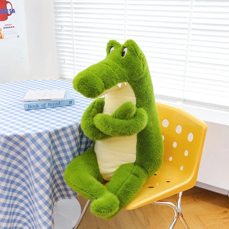 Peluche de cocodrilo verde sentado en posición vertical junto a una mesa con brazos cruzados y un hocico largo
