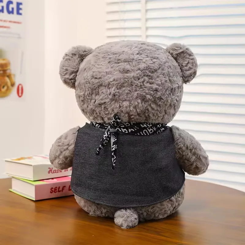 Vista trasera del peluche de oso teddy gris mostrando nudo de bufanda, cobertura del chaleco y pequeno detalle de cola