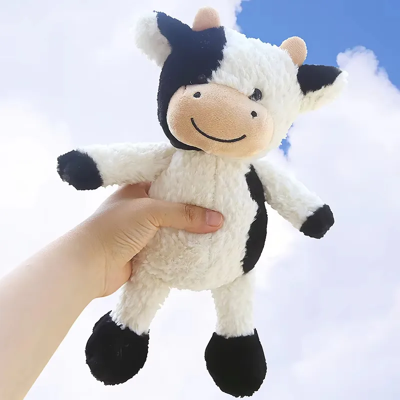 Mano sosteniendo un muñeco de peluche de vaca lechera para mostrar el tamaño del cuerpo suave, las extremidades caídas y los detalles negros de las pezuñas